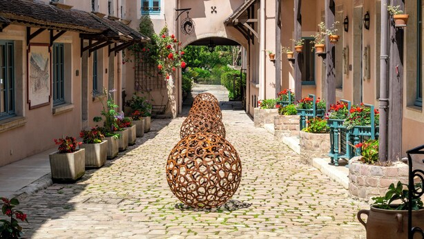 A cobblestone street flanked by buildings with flower boxes and a large decorative metal sculpture.