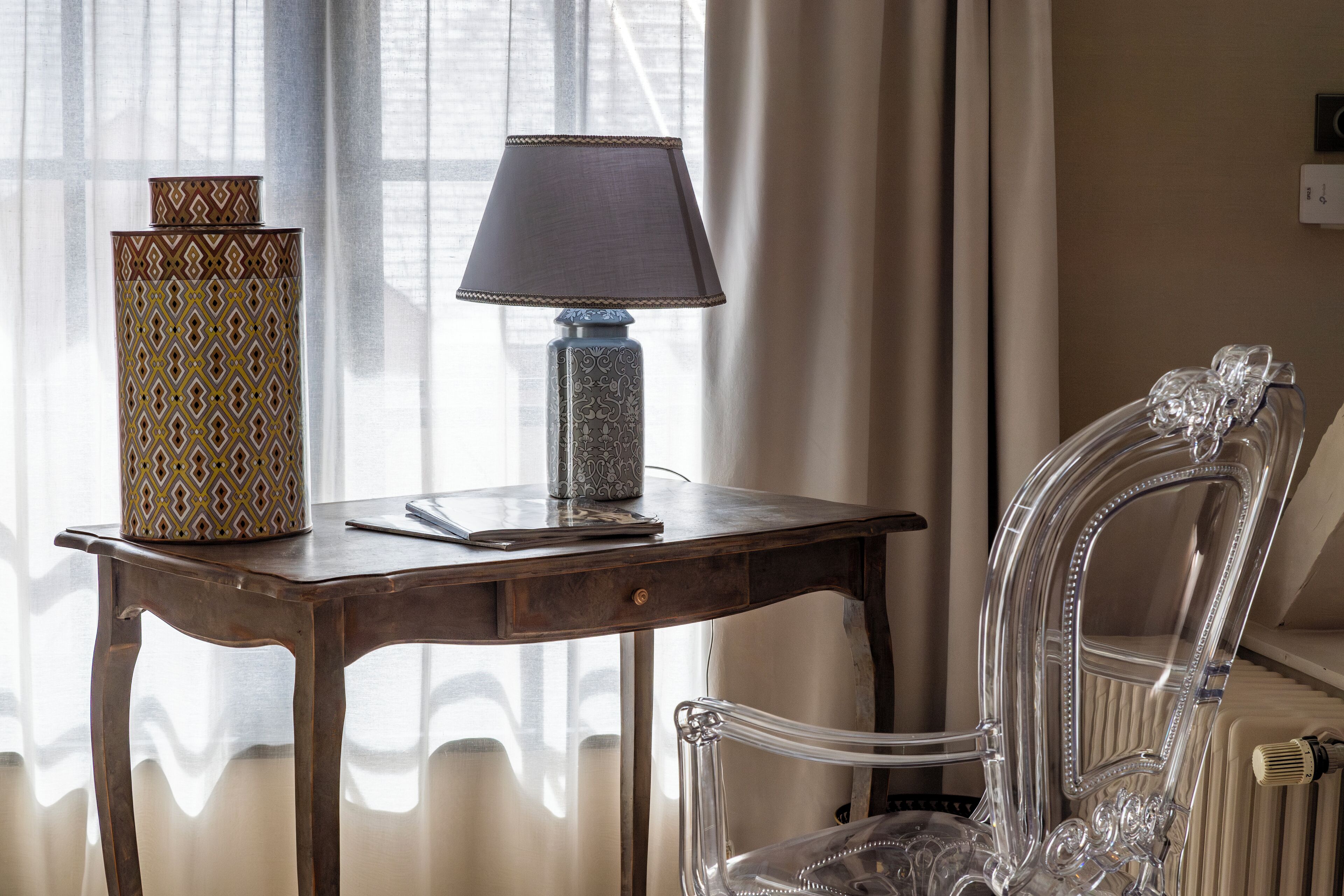 A wooden table with a patterned vase and a lamp, a clear chair, and a window with curtains.