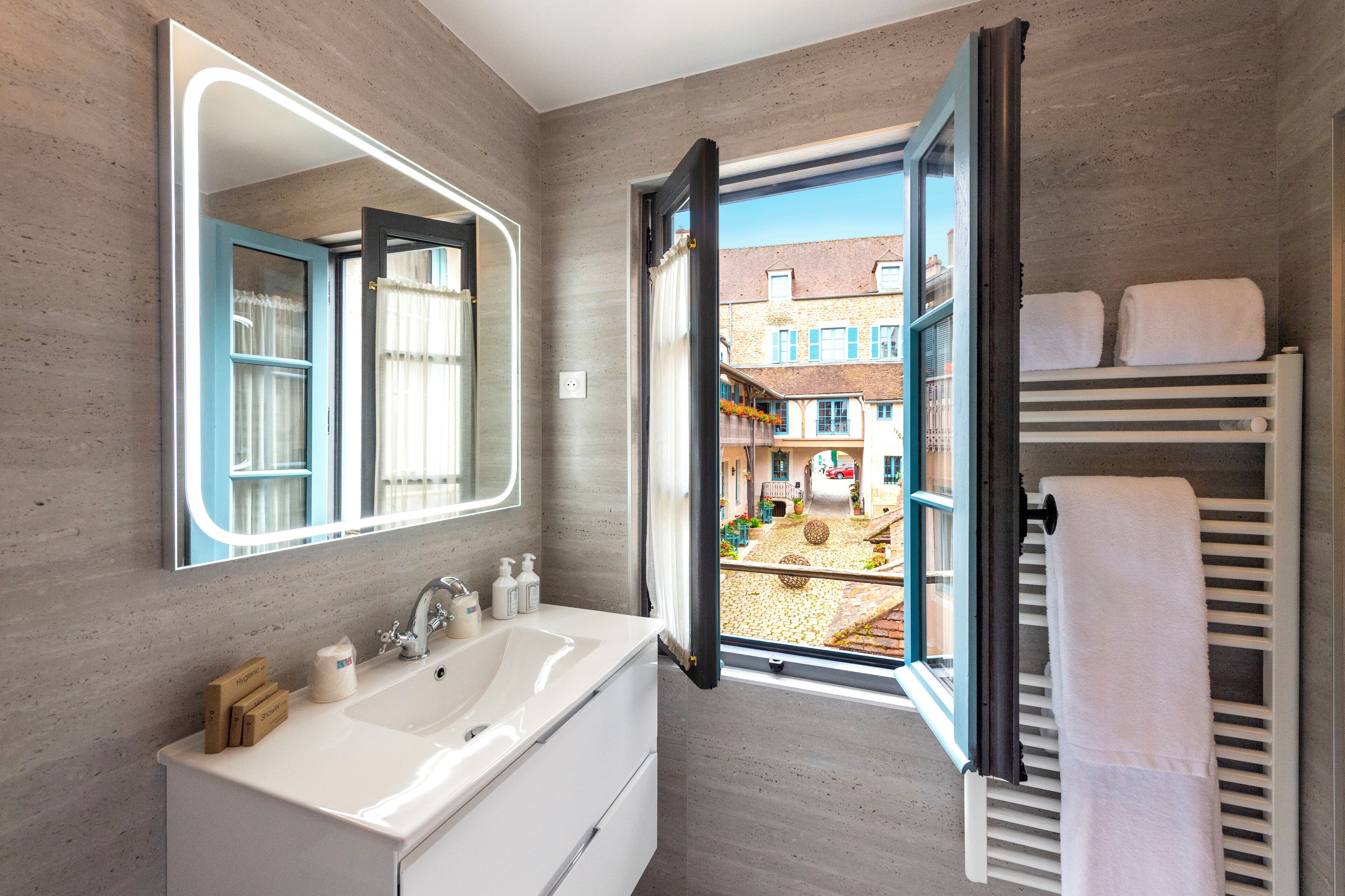 Un baño moderno con un espejo grande, un lavamanos blanco y una ventana que ofrece vistas abiertas a un patio.