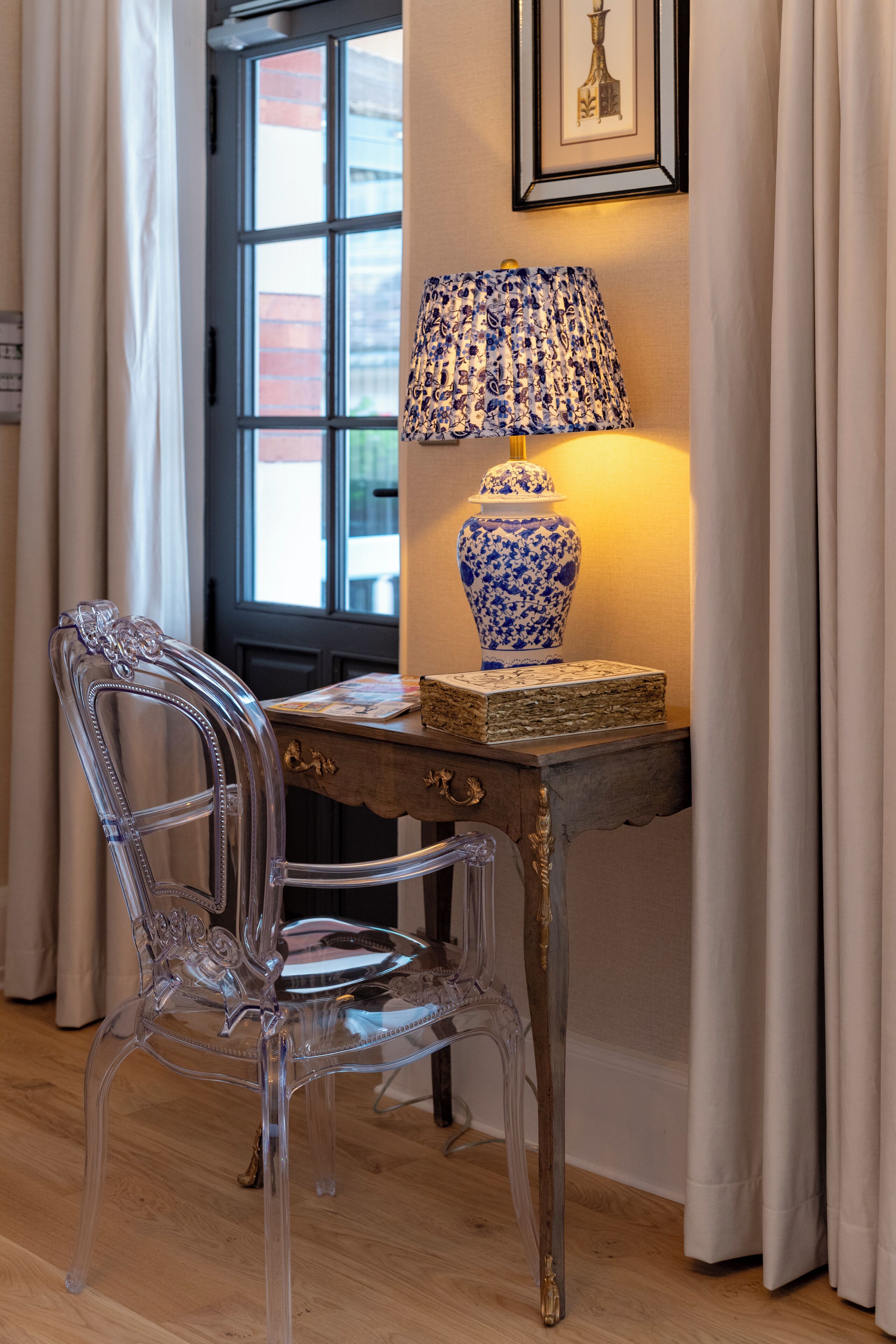 A clear glass chair, a wooden table with a blue and white patterned lamp, and a framed picture on the wall.
