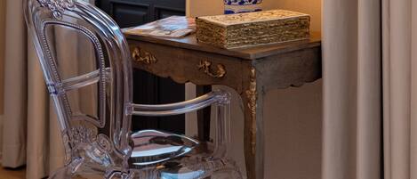 A clear glass chair, a wooden table with a blue and white patterned lamp, and a framed picture on the wall.