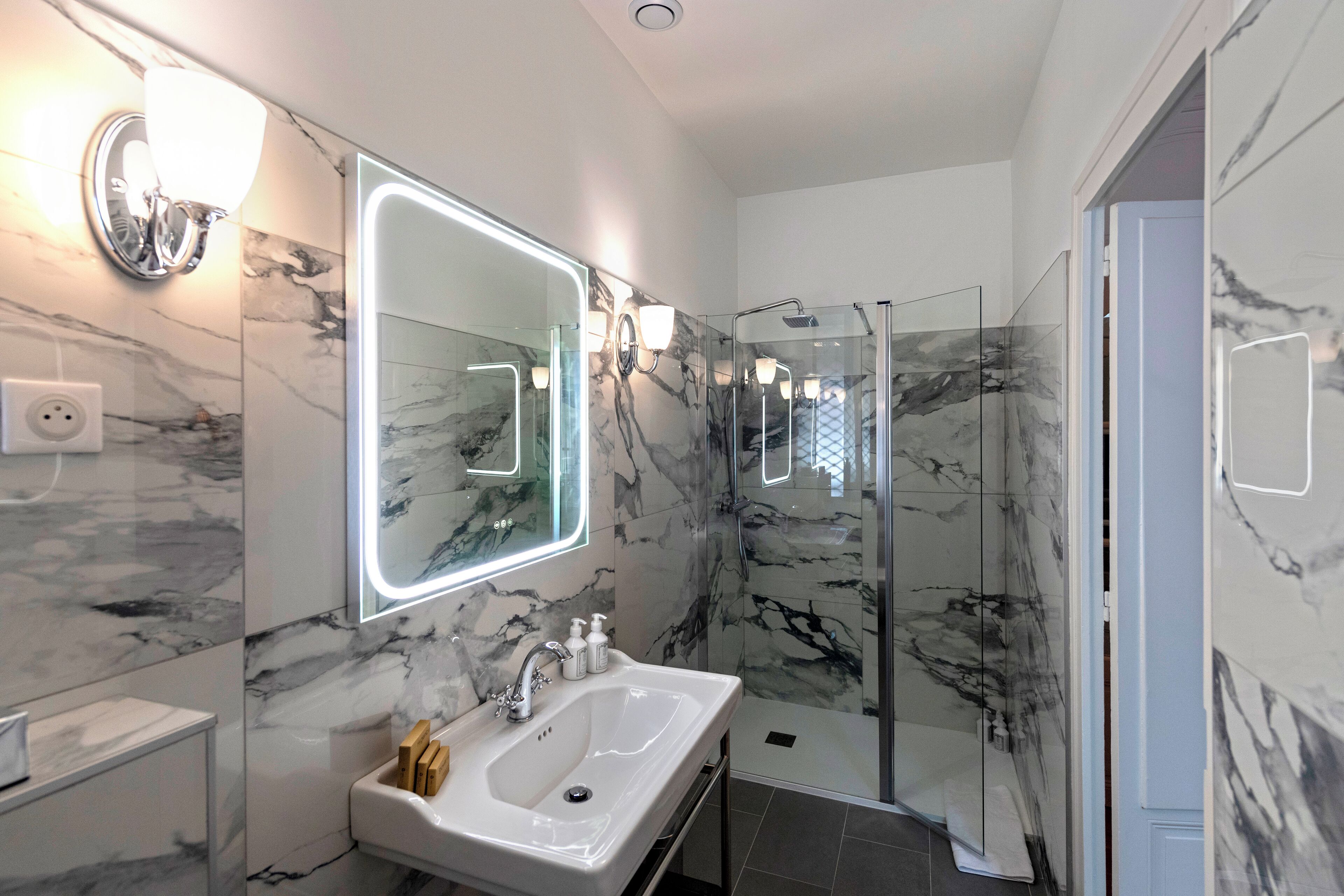 A modern bathroom with a marble wall, a glass-enclosed shower, and a large mirror with integrated lighting.