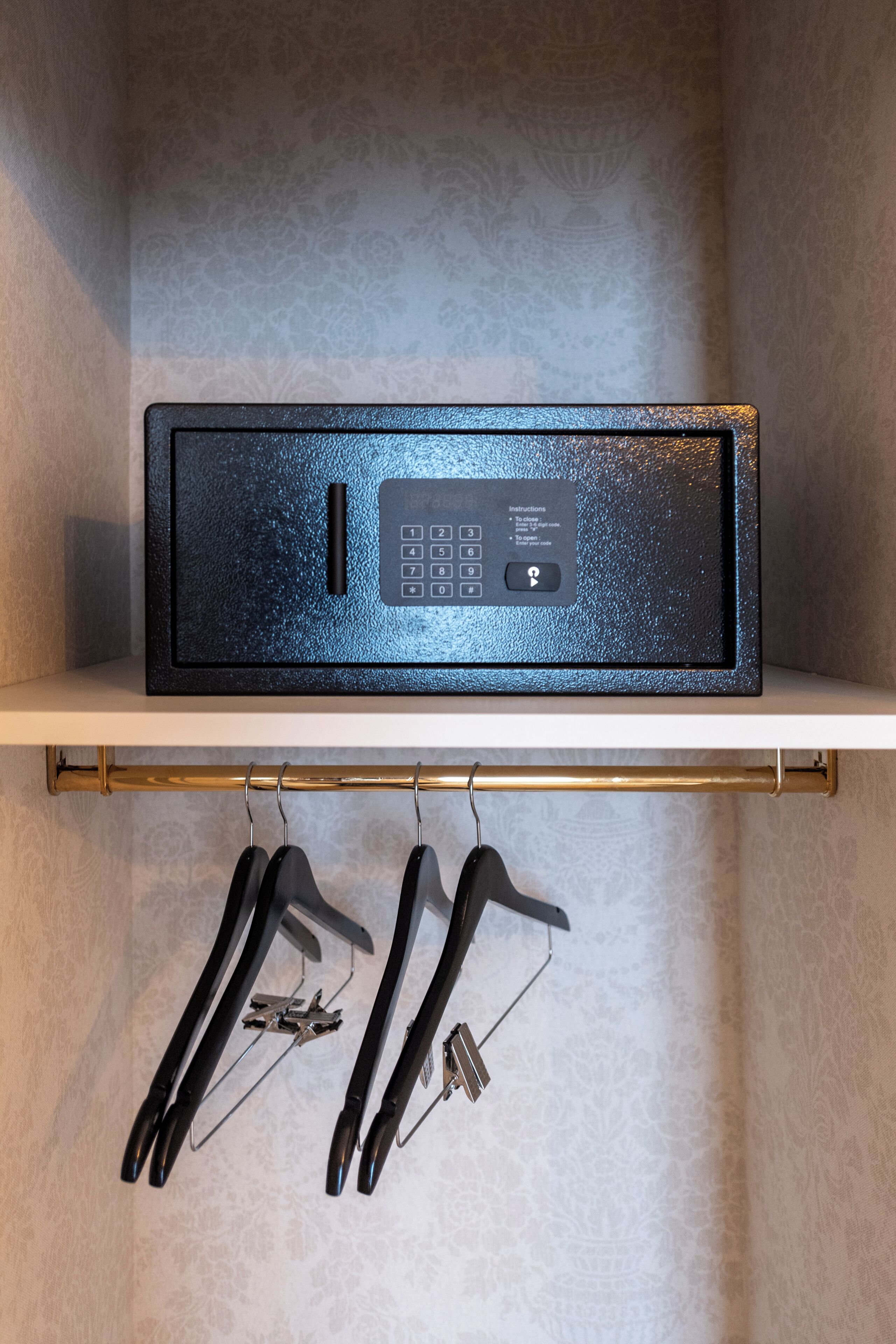 A black safe with a keypad on a white shelf, and black hangers with clothes below.