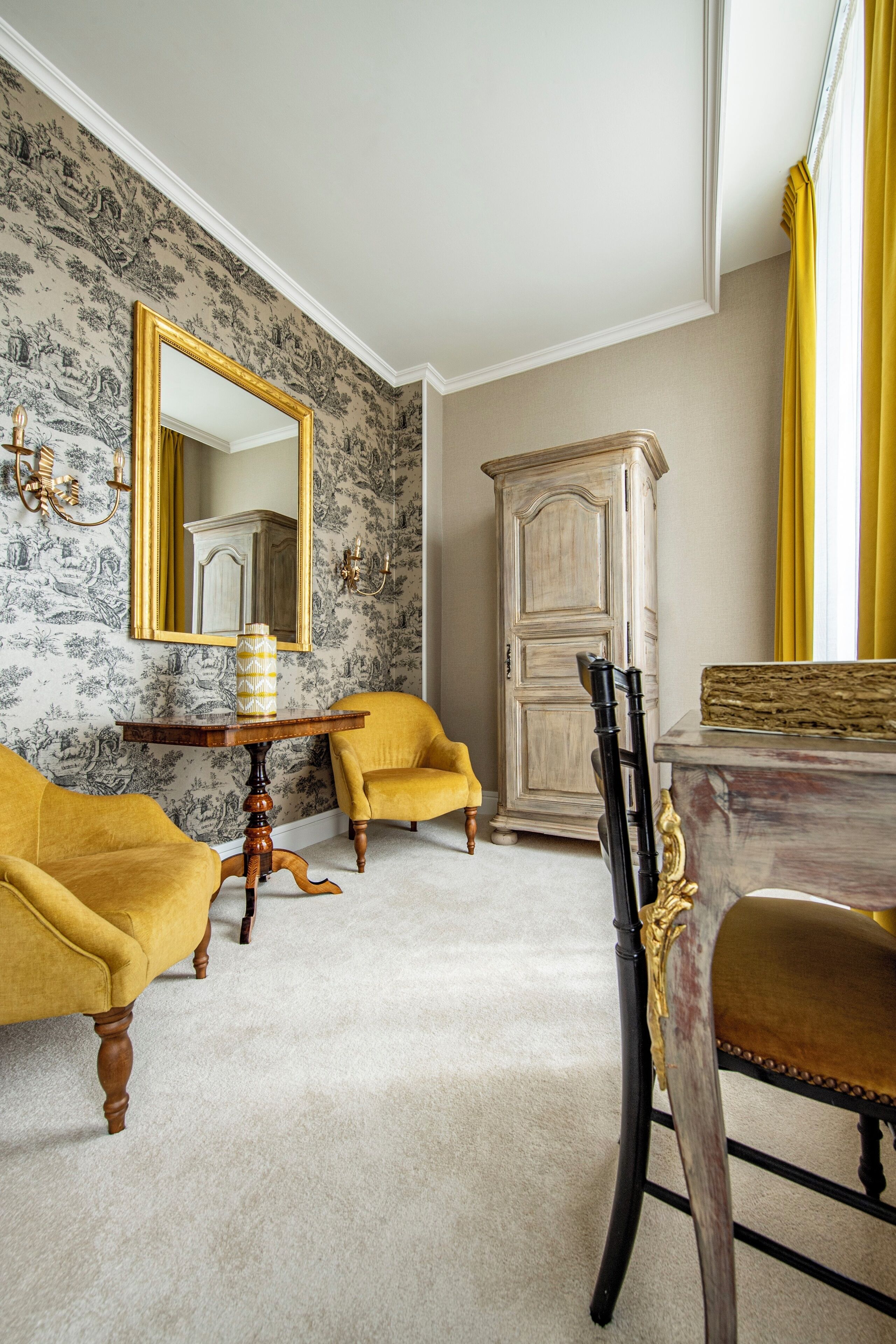 A room with a floral wallpaper, two yellow armchairs, a wooden cabinet, and a dining table with chairs.