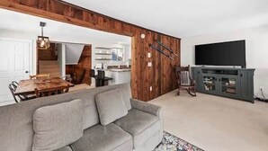 Ein Wohnzimmer mit grauem Sofa, Holzvertäfelung, einem Fernseher auf einem Schrank und einem Essbereich mit Holztisch und Stühlen.