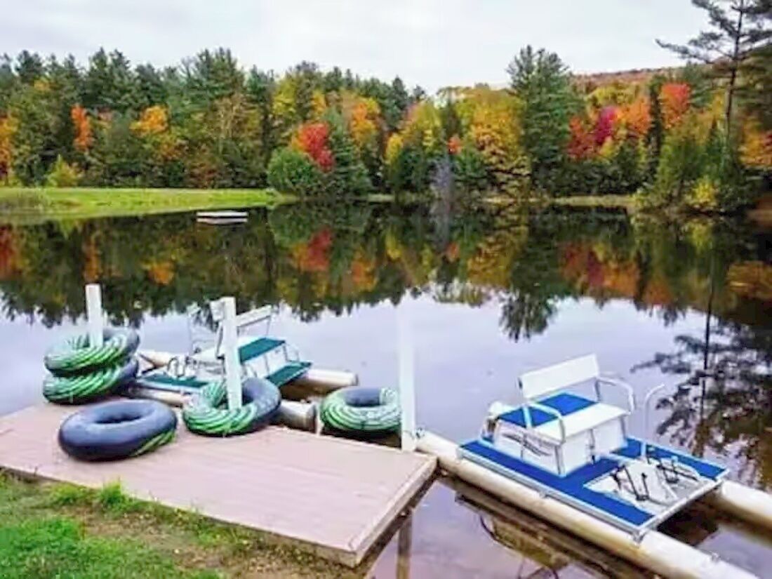 Un muelle con un bote y anillos inflables, rodeado por un lago tranquilo y árboles de colores otoñales.