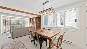 Ein geräumiger Wohnbereich mit einem Holzessttisch, schwarzen Stühlen, einem grauen Sofa und einem großen Fenster mit Jalousien.