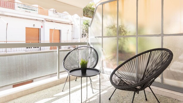 A balcony with two black wicker chairs and a small table, a large window with frosted glass panels, and a view of a building with a closed shutter. - Petite 1BR Apartment in Exarchia (Athens)