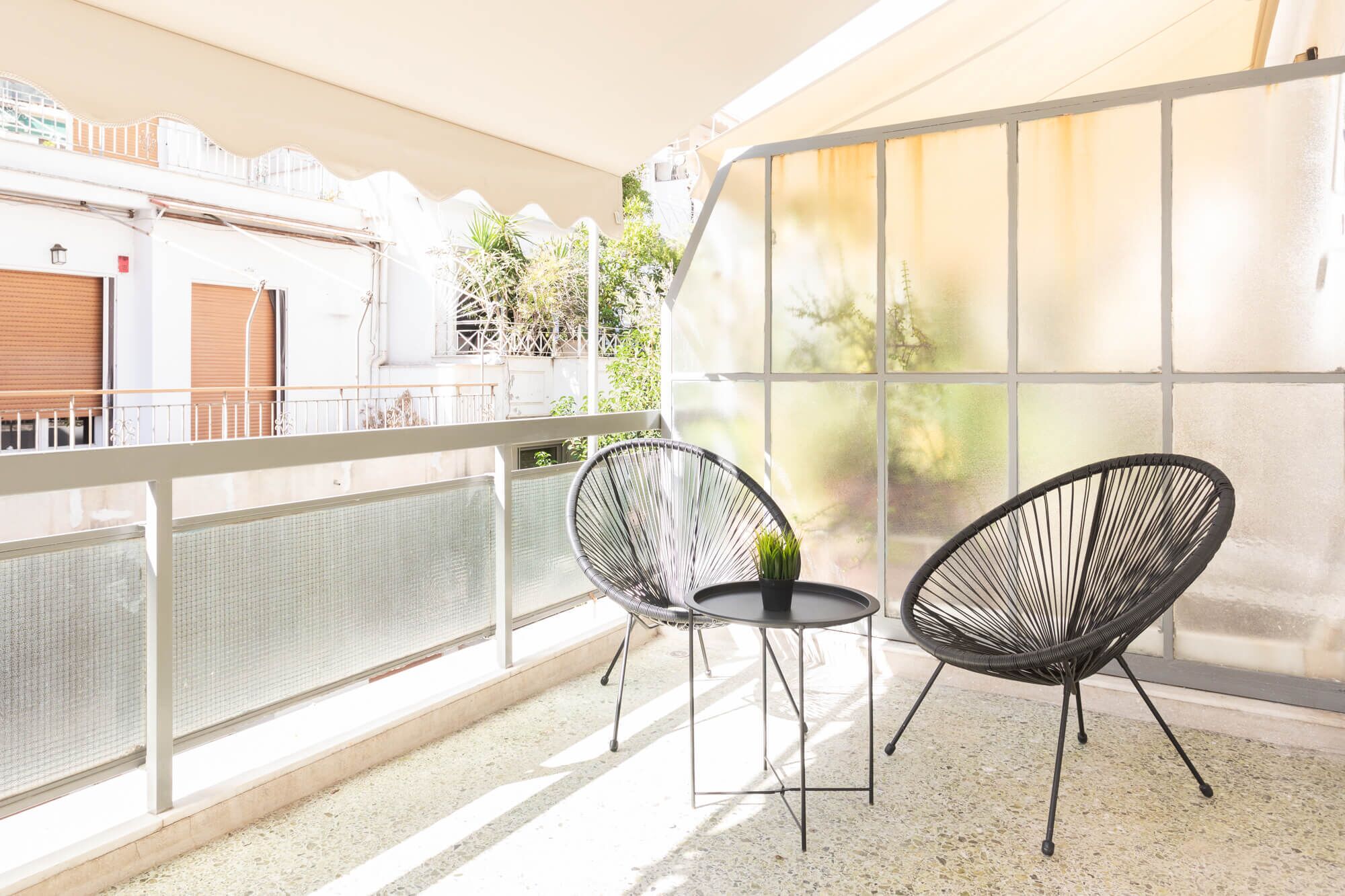 A balcony with two black wire chairs and a small table, overlooking a street with buildings and greenery.