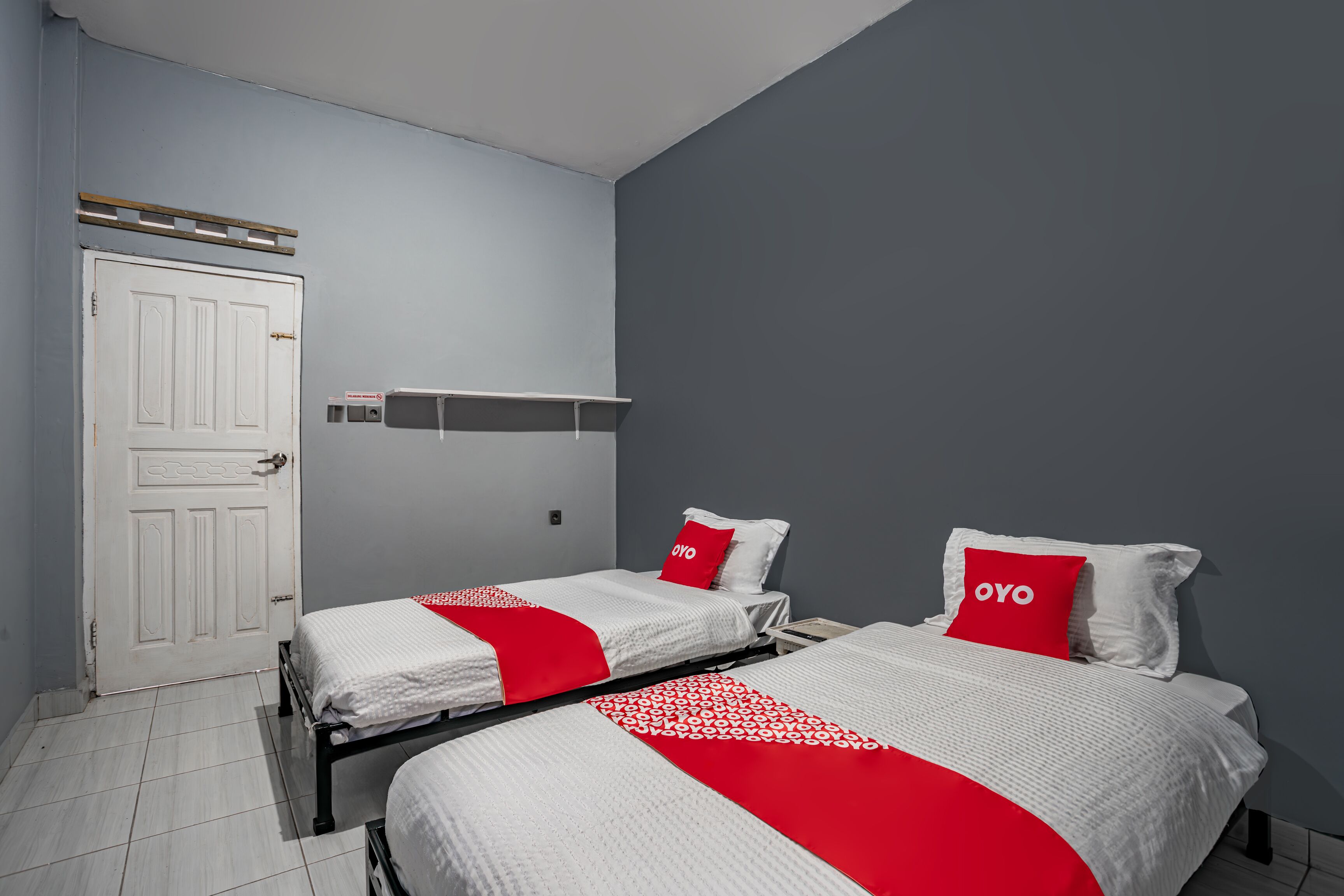 Ett hotellrum med två sängar, var och en med ett rött och vitt sängöverkast och matchande kuddar. Det finns en vit dörr och en hylla ovanför.