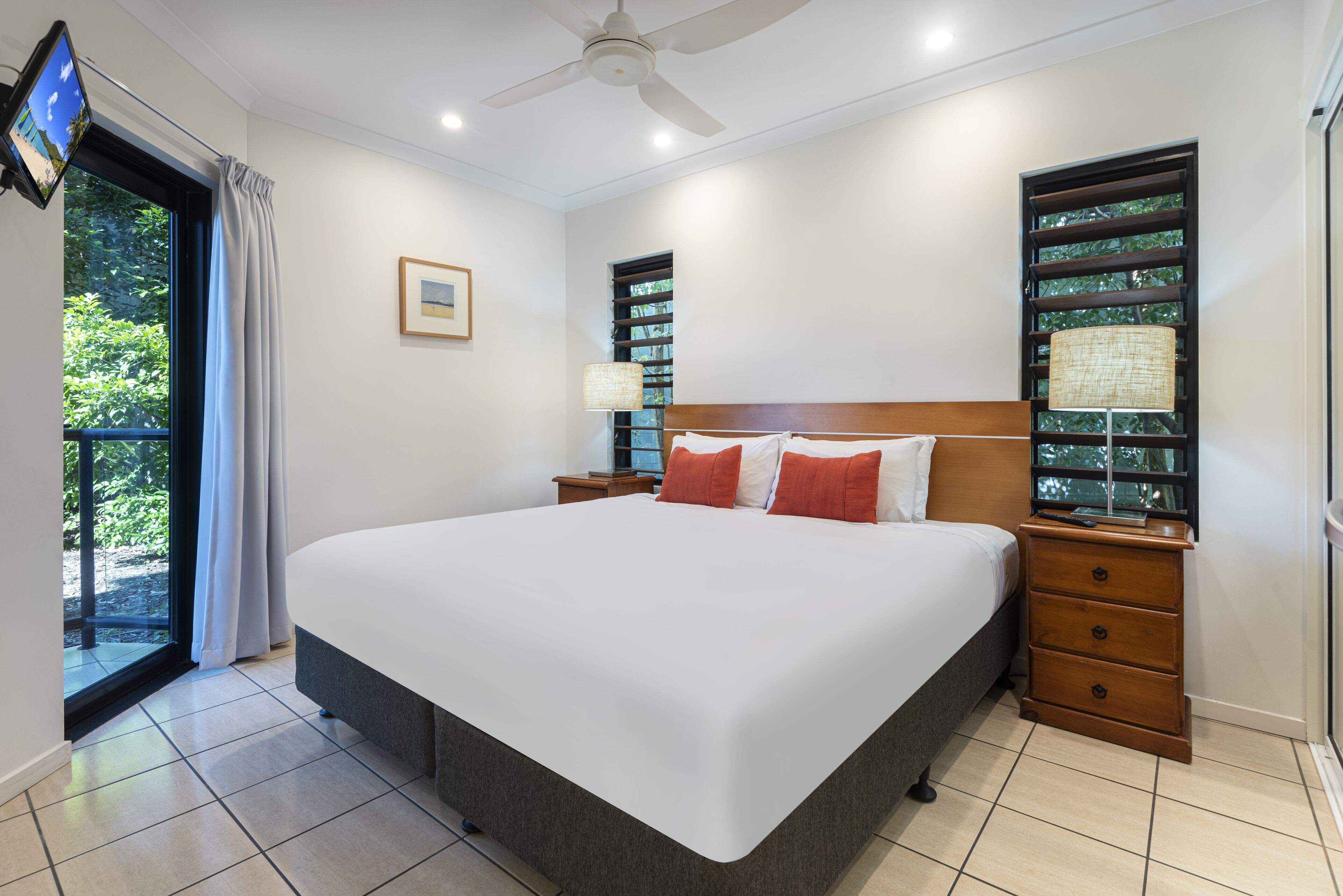 A bedroom with a large bed, a ceiling fan, a TV mounted on the wall, and a window with curtains.