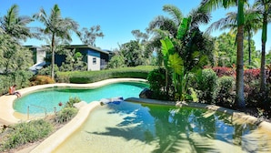 Une piscine entourée d’une végétation tropicale, avec un bâtiment en arrière-plan.
