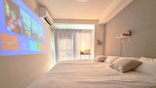 A bedroom with a bed, a projector screen displaying movie, music, and photo icons, and a window with curtains. - City Heights (Osaka)