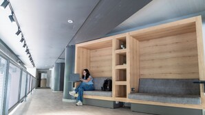 A modern waiting area with wooden benches and a grey couch, a laptop on the bench, and a backpack on the couch. - Athens Hawks Urban (Athens)
