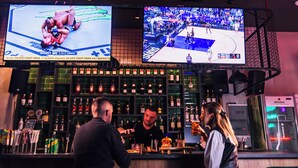 A bar area with multiple screens displaying sports events, a bartender, and a customer. - Athens Hawks Urban (Athens)