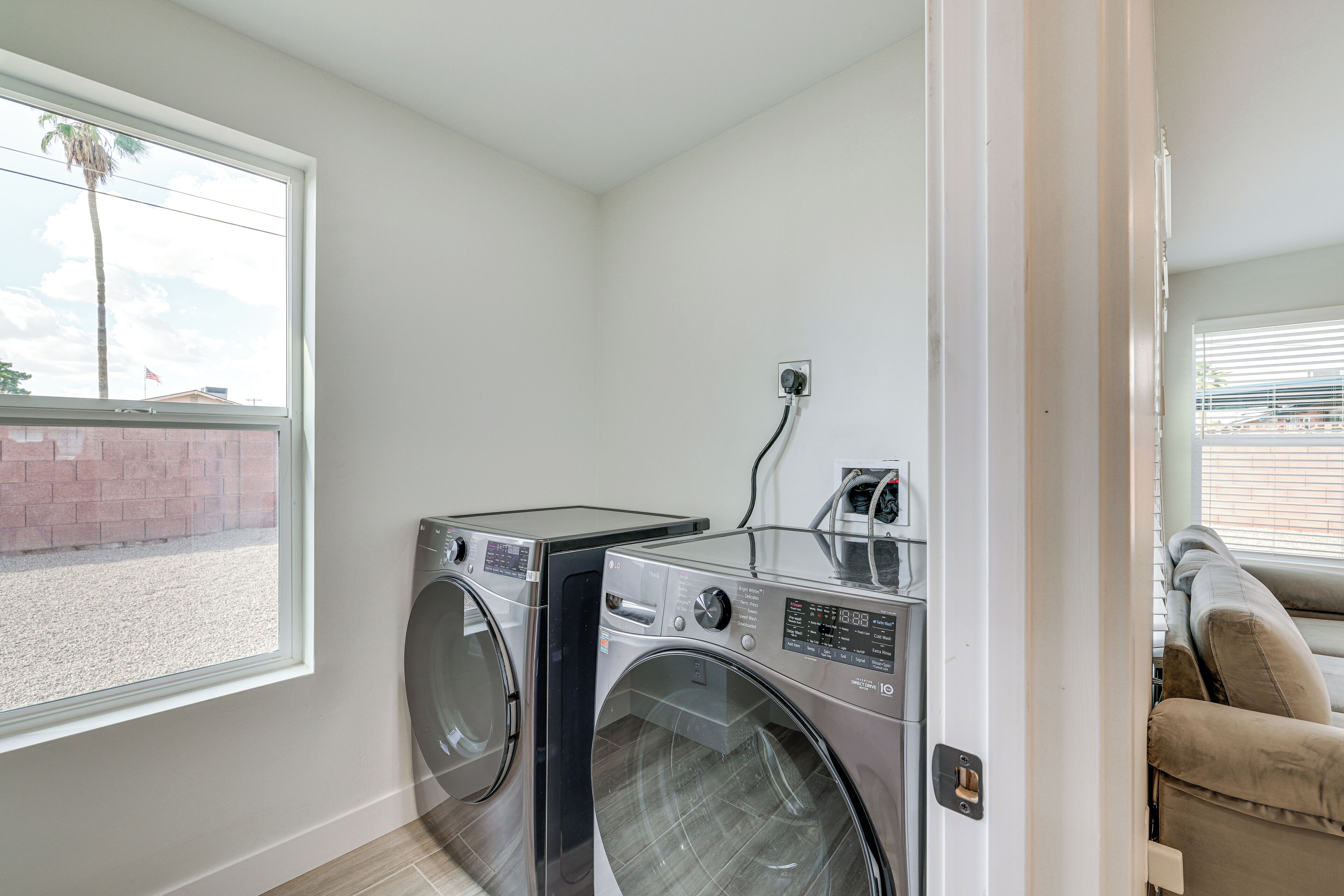 A laundry room with two front-loading washing machines and a dryer, a window with a view of a palm tree and a brick wall, and a door leading to a living area with a beige sofa.