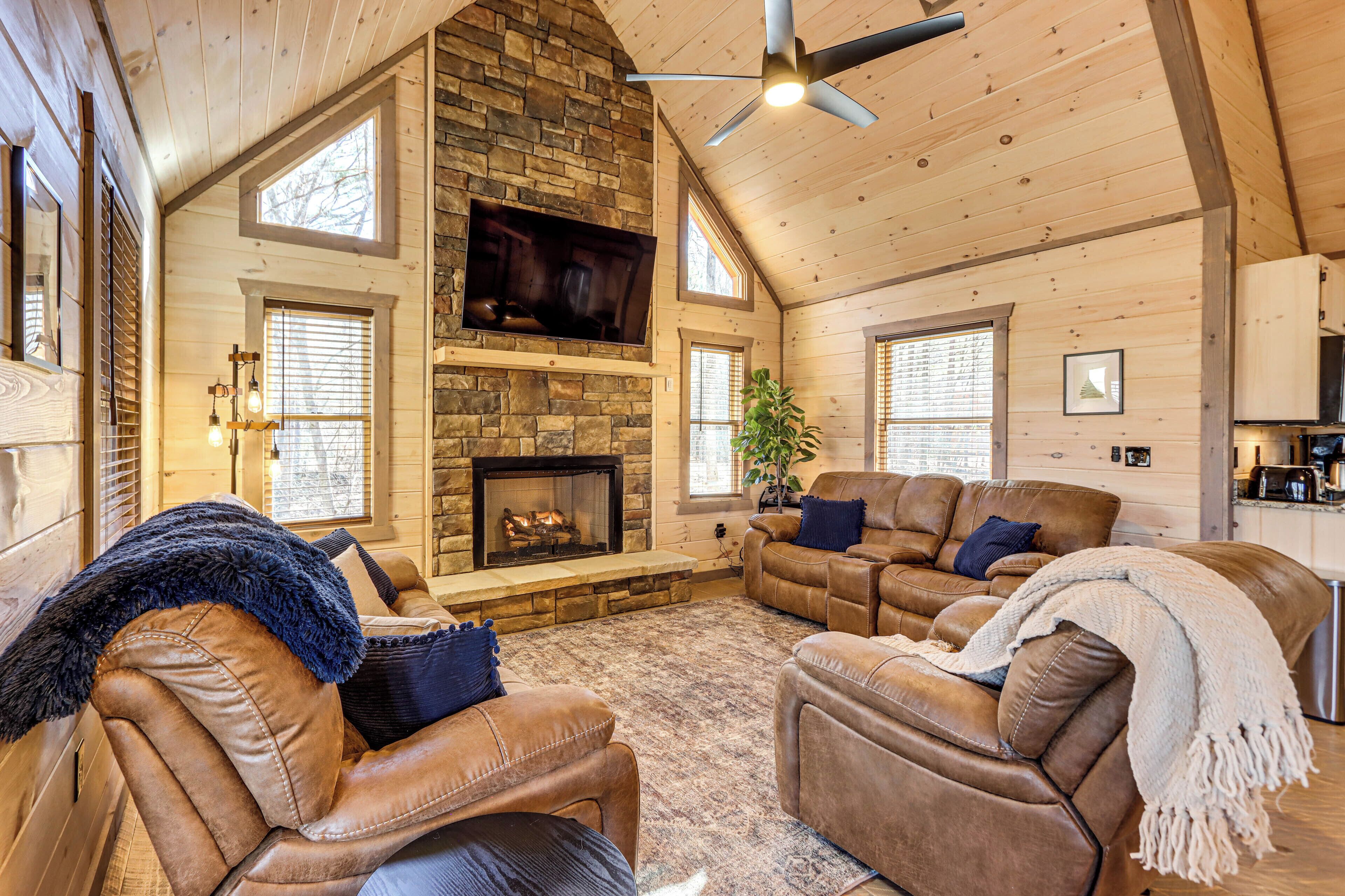 A cozy log cabin interior with a stone fireplace, leather sofas, and a television mounted above it.