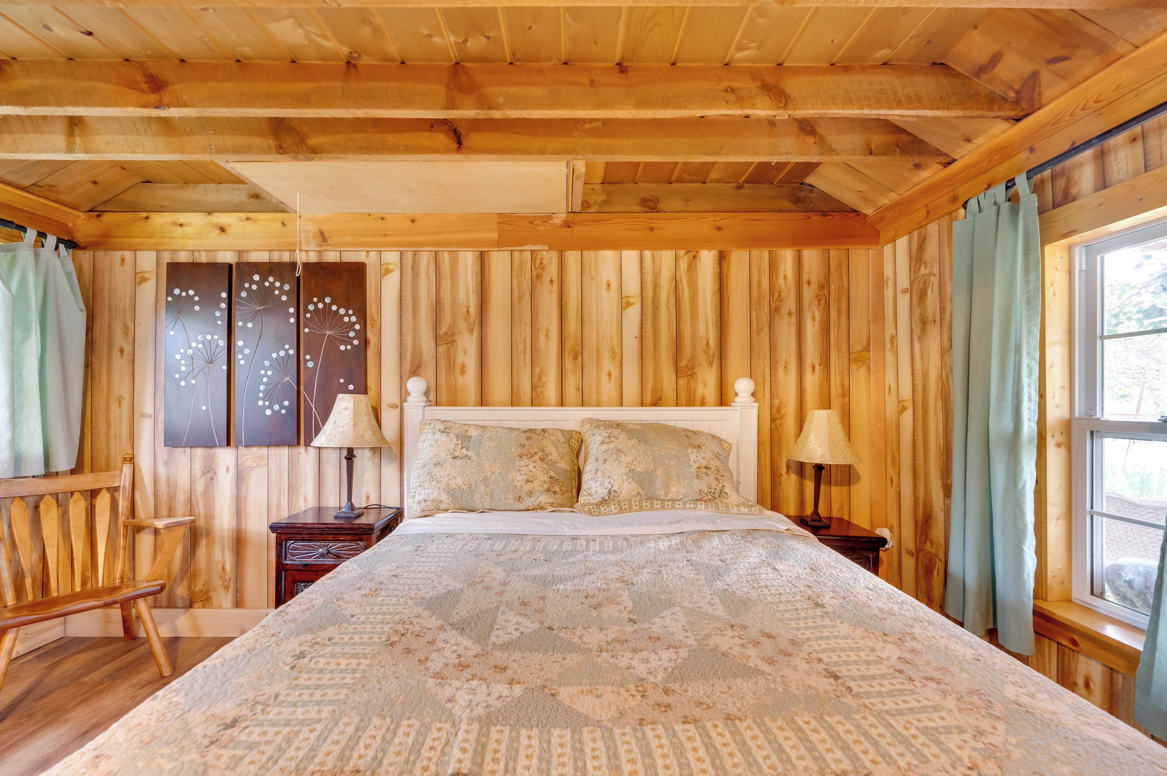A wooden cabin bedroom with a bed, two nightstands, a chair, and a window with curtains.
