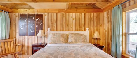 A wooden cabin bedroom with a bed, two nightstands, a chair, and a window with curtains.