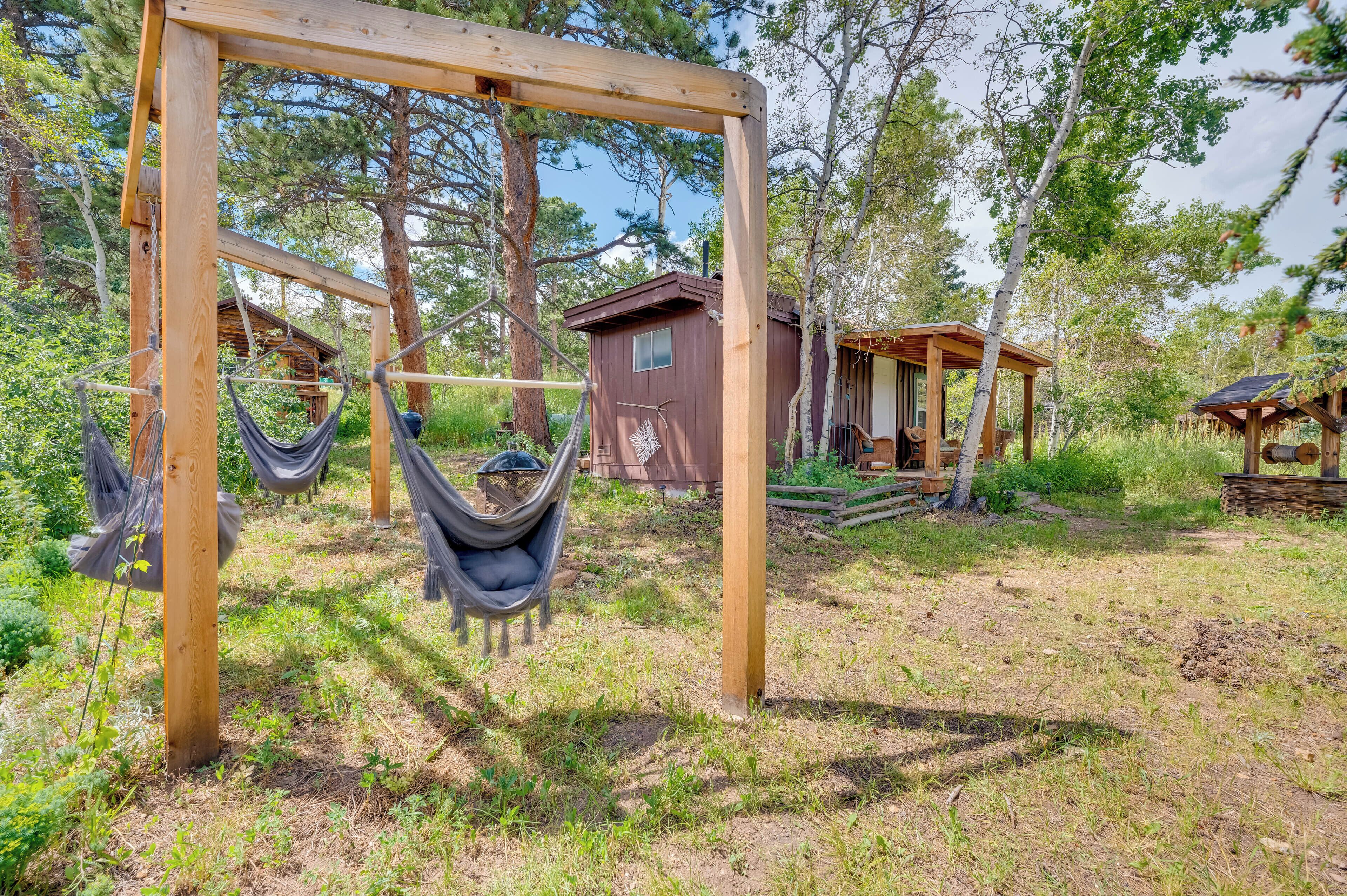 A wooden swing structure with a hammock, a small wooden cabin, and a thatched-roof building in a forested area.