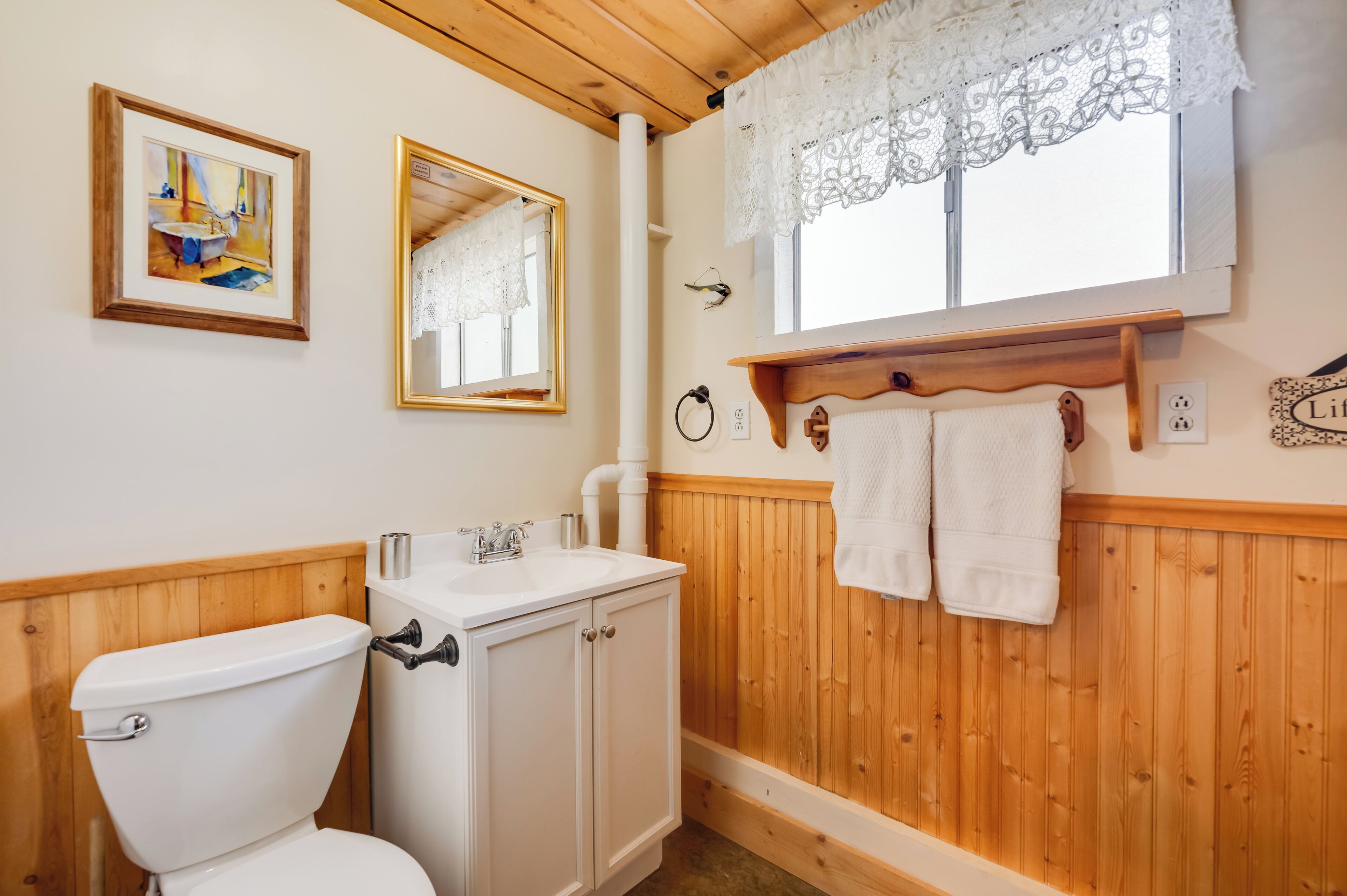 A bathroom with a wooden vanity, a toilet, a sink, and a mirror. There is a window with lace curtains and a towel rack with towels.
