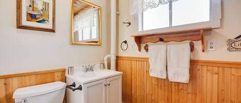 A bathroom with a wooden vanity, a toilet, a sink, and a mirror. There is a window with lace curtains and a towel rack with towels.