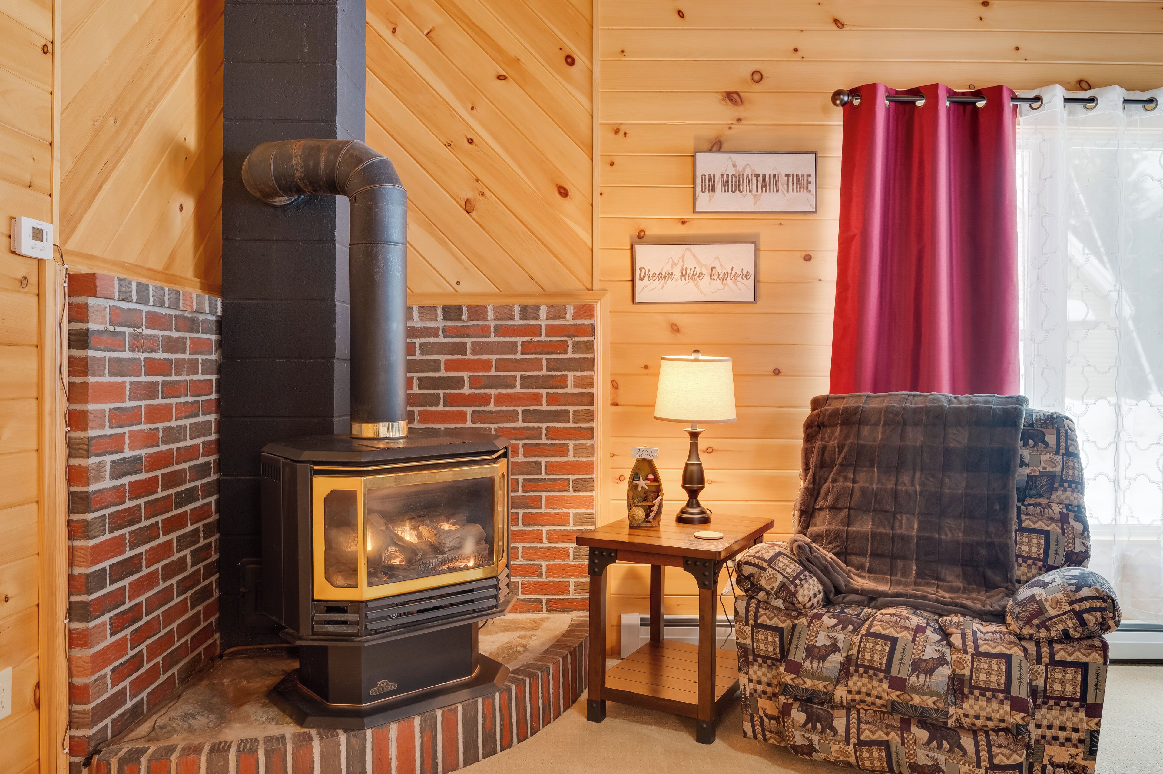 A cozy room with a brick fireplace, a wooden ceiling, a patterned armchair, and a small table with a lamp.