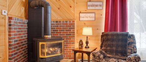 A cozy room with a brick fireplace, a wooden ceiling, a patterned armchair, and a small table with a lamp.