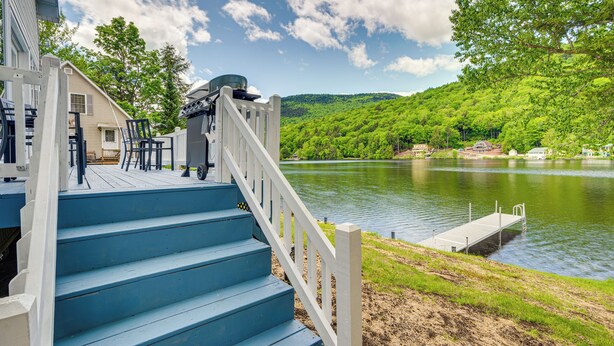 Una terraza de madera con escalones azules que conducen a un muelle junto a un lago, rodeado de vegetación y casas.