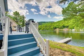Une terrasse en bois avec des marches bleues menant à un quai au bord d’un lac, le tout entouré de verdure et de maisons.