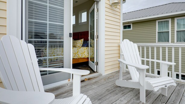 A wooden deck with two white Adirondack chairs, a sliding glass door leading to a bedroom with a bed and a window, and a green building in the background.