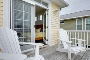 A wooden deck with two white Adirondack chairs, a sliding glass door leading to a bedroom with a bed and a window, and a green building in the background. - Carolina Beach Home w/ Balconies, Walk to Beach! (Carolina Beach)