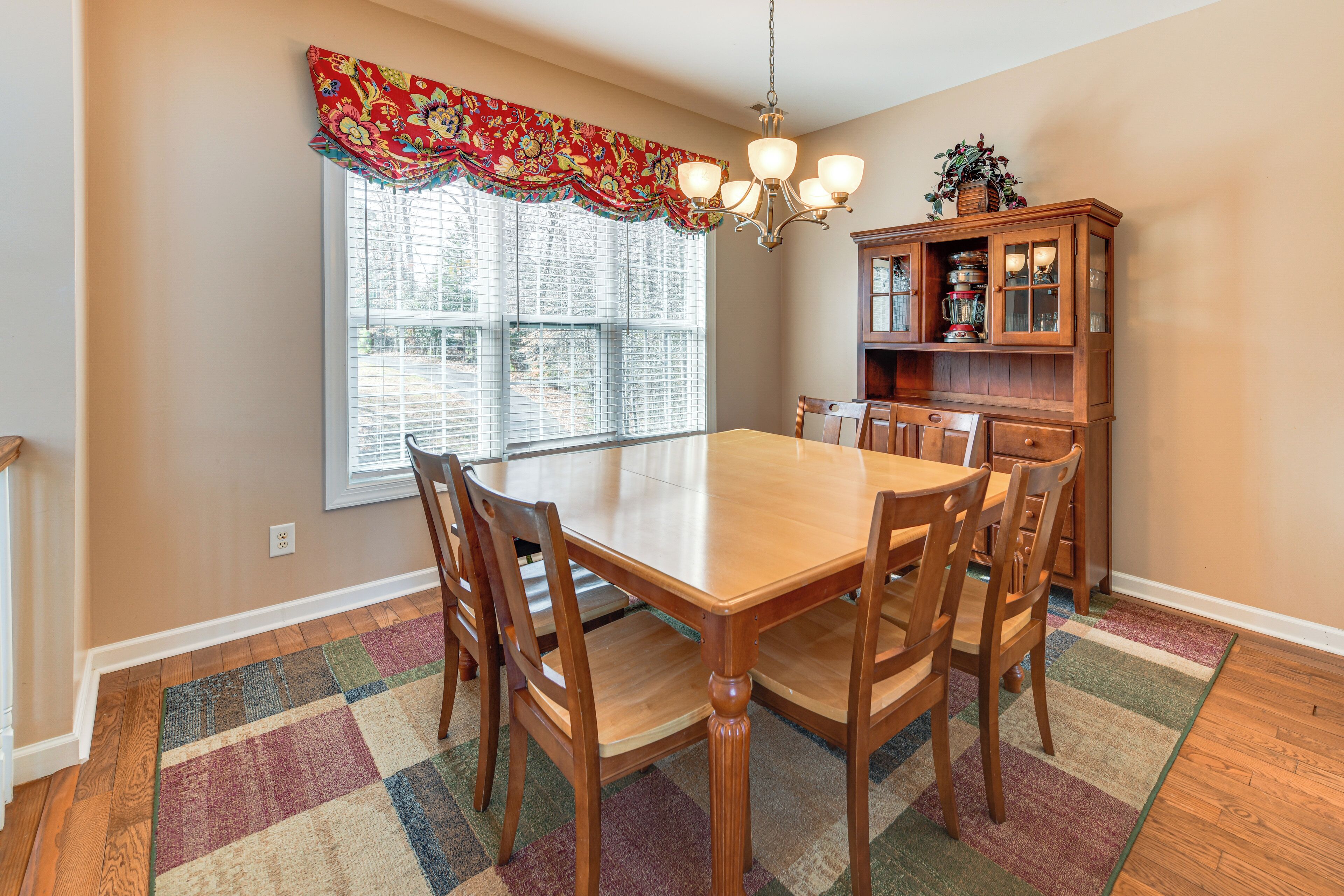 A dining room with a wooden table and chairs, a window with blinds, a chandelier, and a wooden cabinet with glass doors.