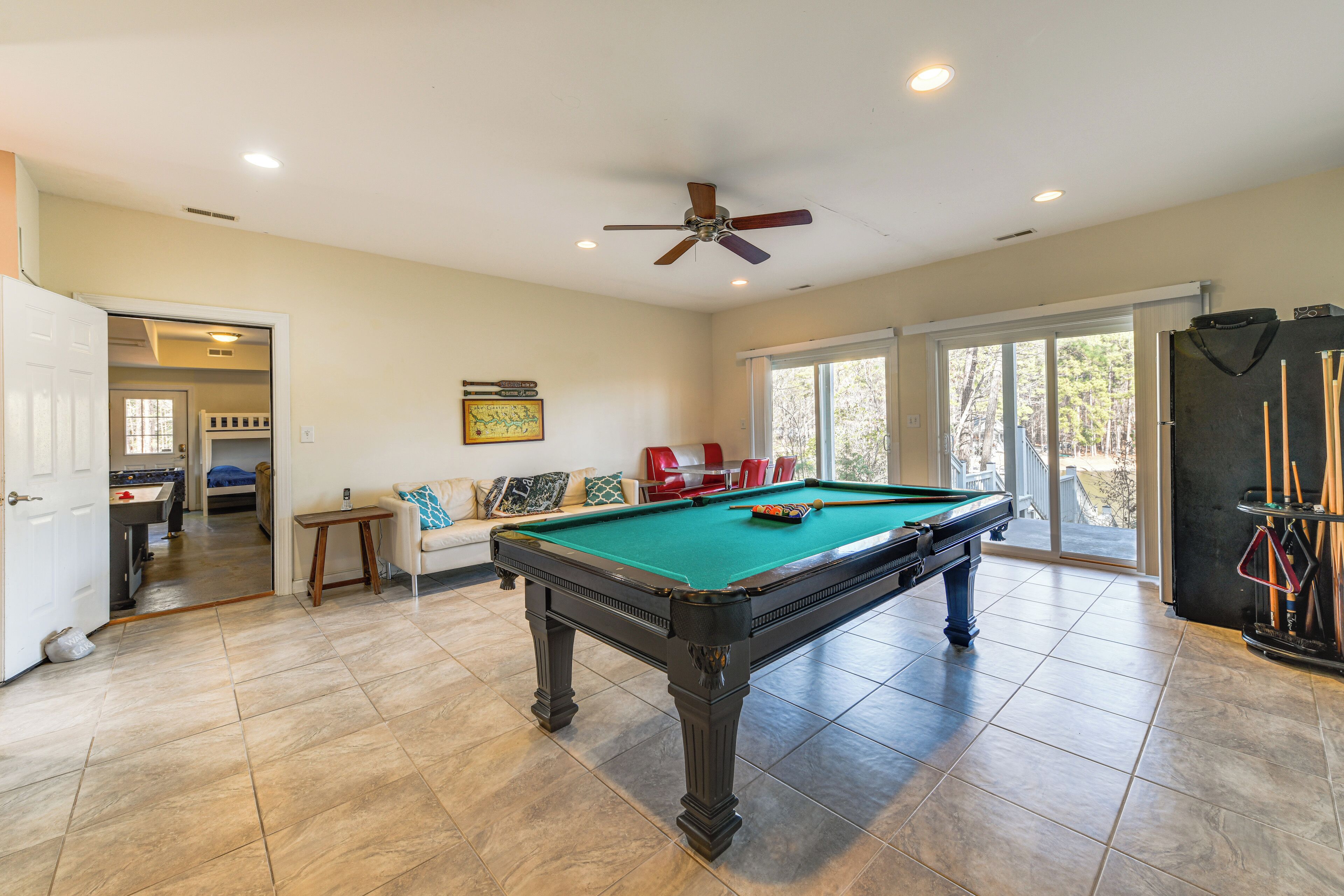 A pool table in a well-lit room with a ceiling fan, a sofa with cushions, and a door leading to another room.