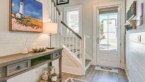 A hallway with a staircase, a painting of a lighthouse, a lamp, and a door leading outside. - Stunning Santa Rosa Beach Home, 1/2 Mi to Ocean! (Santa Rosa Beach)