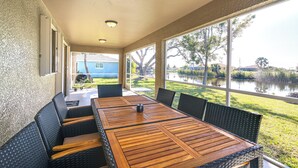 A covered patio with a wooden table and chairs, large windows, and a view of a lake and greenery. - Canal-front Cape Coral Home w/ Fire Pit, By Beach! (Cape Coral)