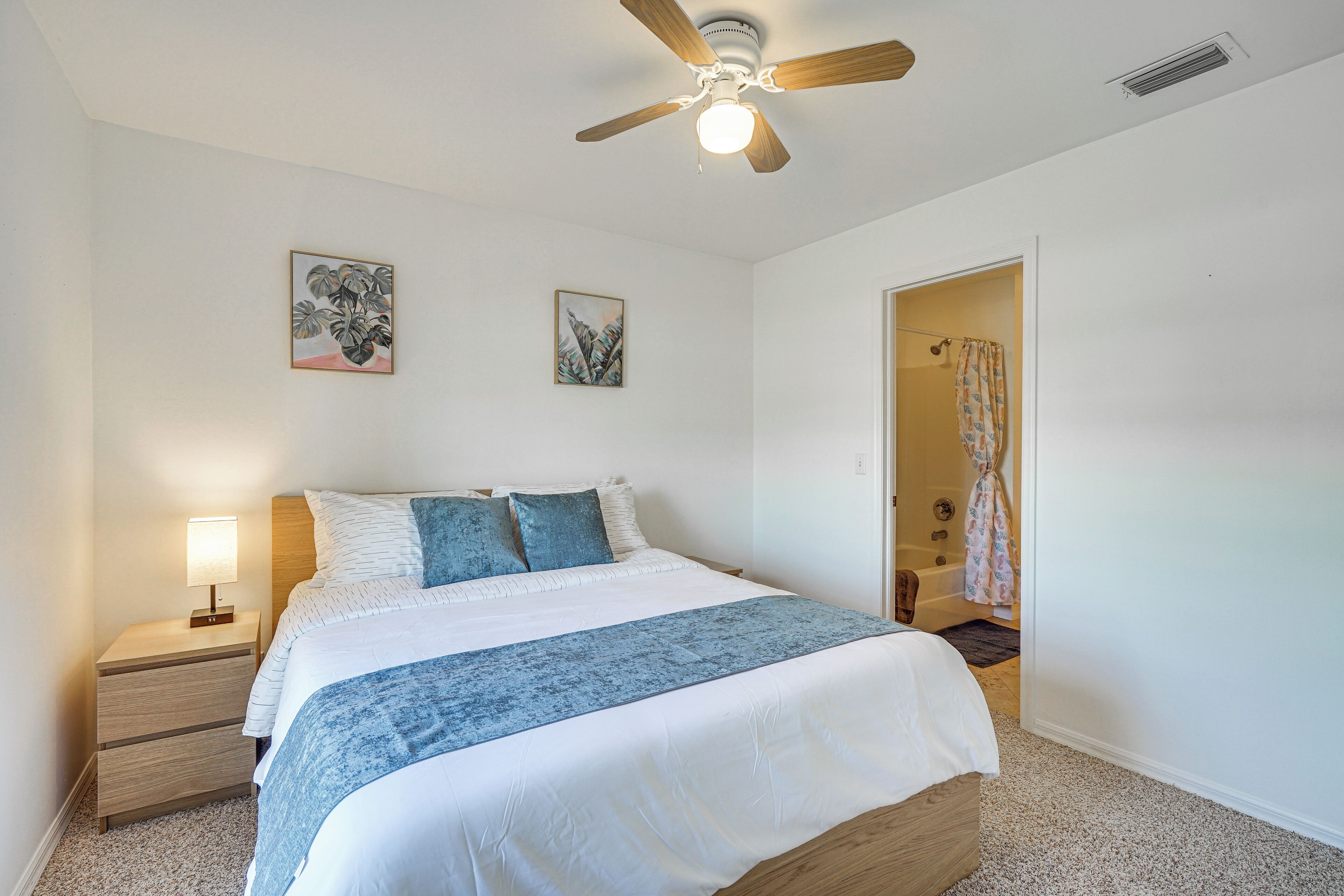 Une chambre à coucher comprenant un lit, des tables de chevet, un ventilateur de plafond et deux œuvres d’art accrochées au mur.