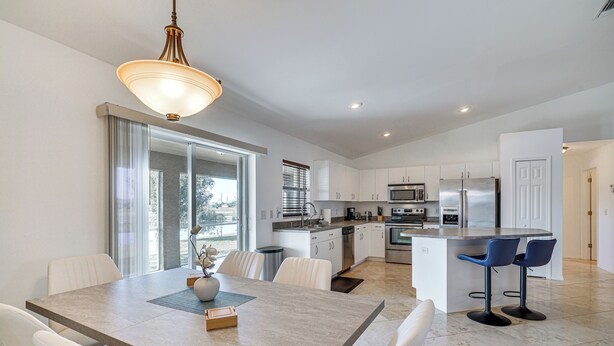 A modern kitchen with white cabinets, a central island with bar stools, and a dining area with a table and chairs. - Canal-front Cape Coral Home w/ Fire Pit, By Beach! (Cape Coral)