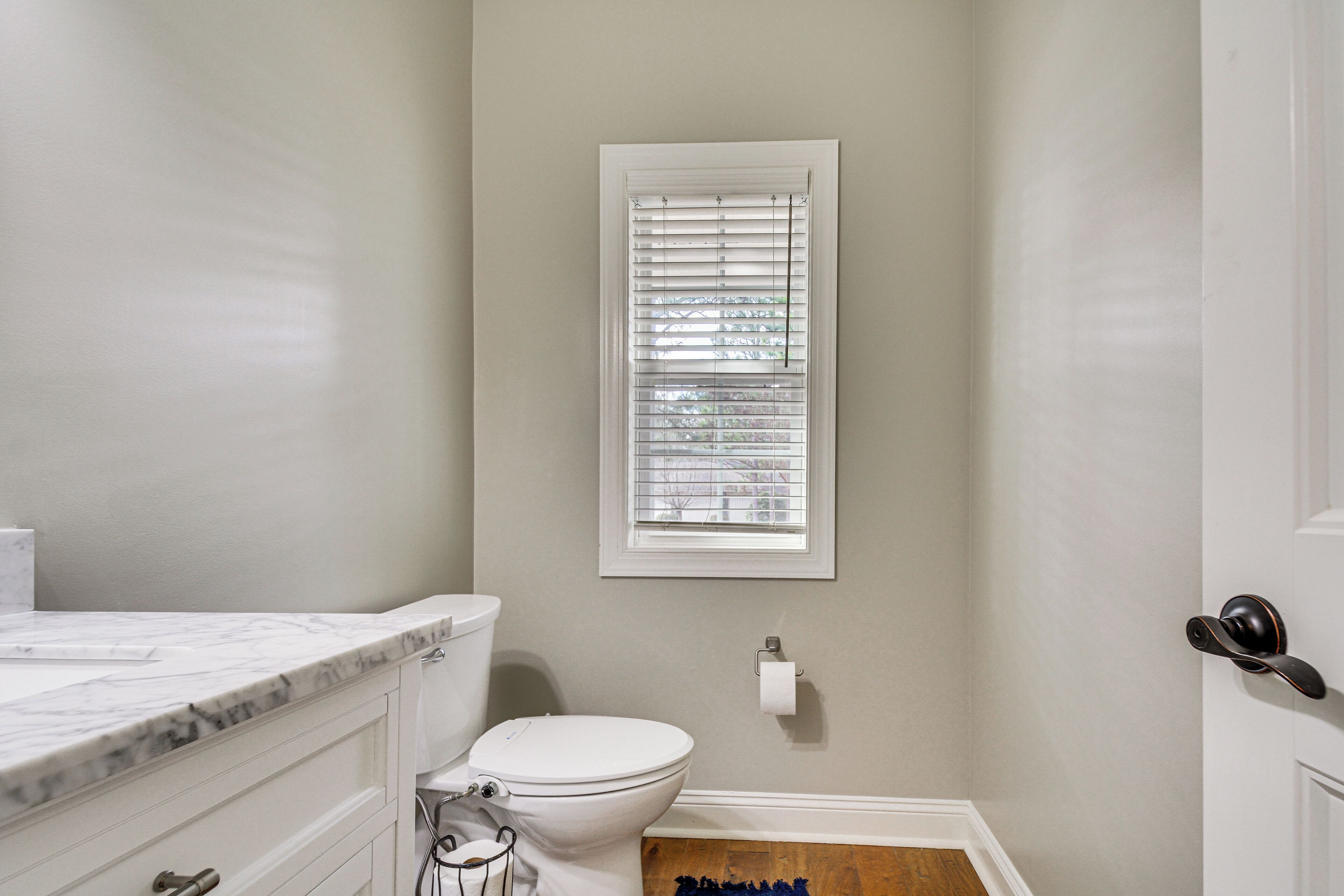A bathroom with a toilet, a marble countertop, a window with blinds, and a door with a round handle.