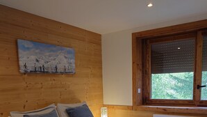Un dormitorio con cabecera de madera, cama con manta a cuadros, mesita de noche con lámpara y ventana con vista a zonas verdes.
