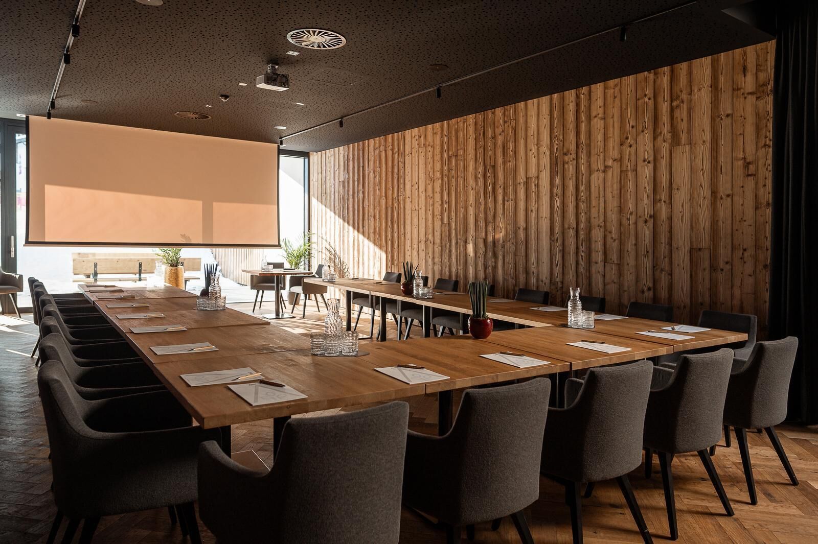 Ein Konferenzraum mit einem langen Holztisch, grauen Stühlen und einer Projektionsleinwand.