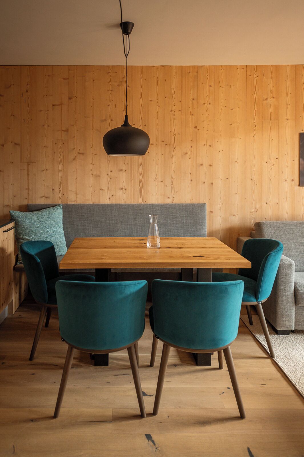 A dining area with a wooden table, teal chairs, a grey sofa, and a hanging light fixture.