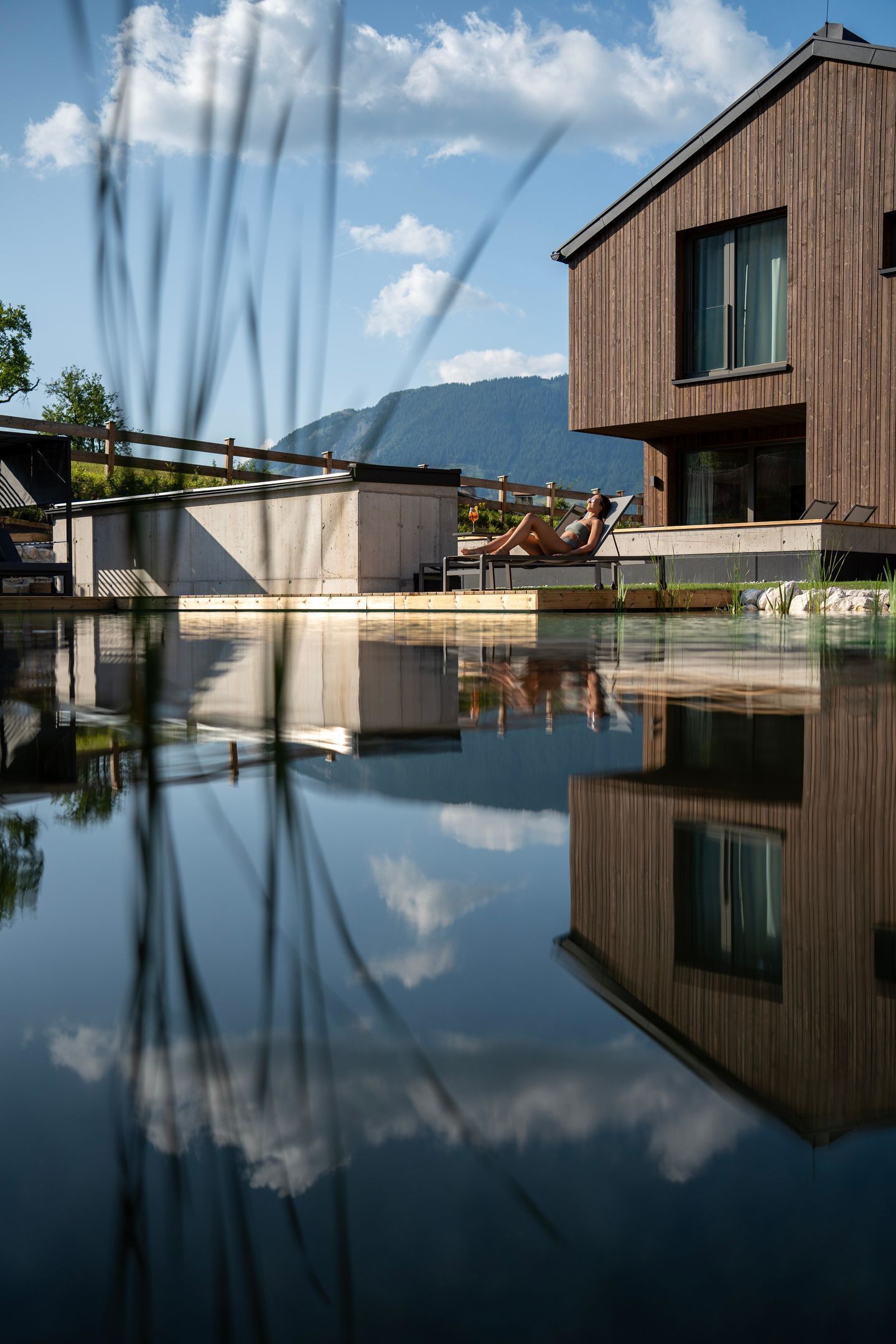A modern building with a pool, a person relaxing by the pool, and mountains in the background.