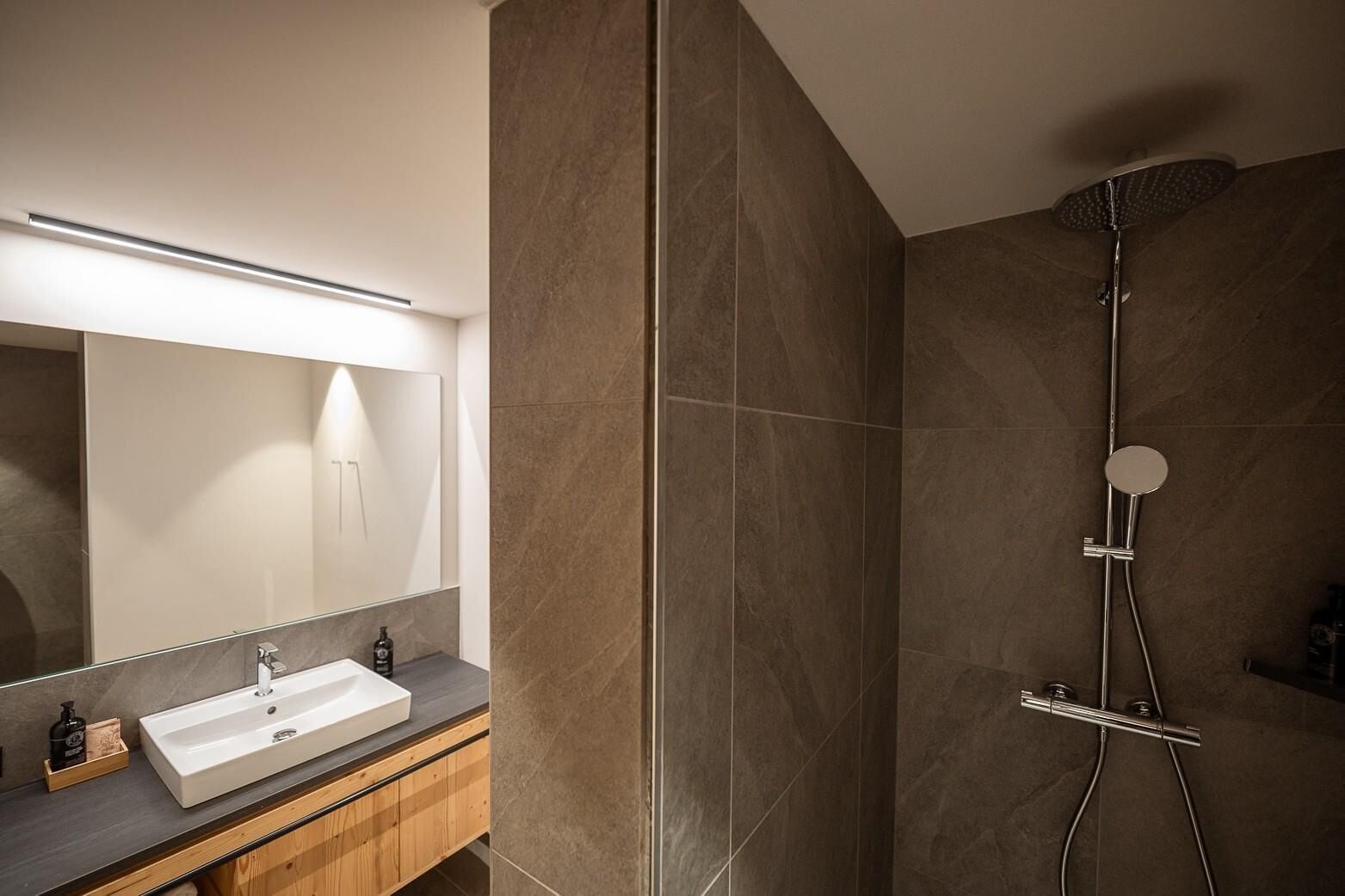 Une salle de bain moderne avec un grand miroir, un lavabo blanc et une douche équipée d’un pommeau de douche à main.