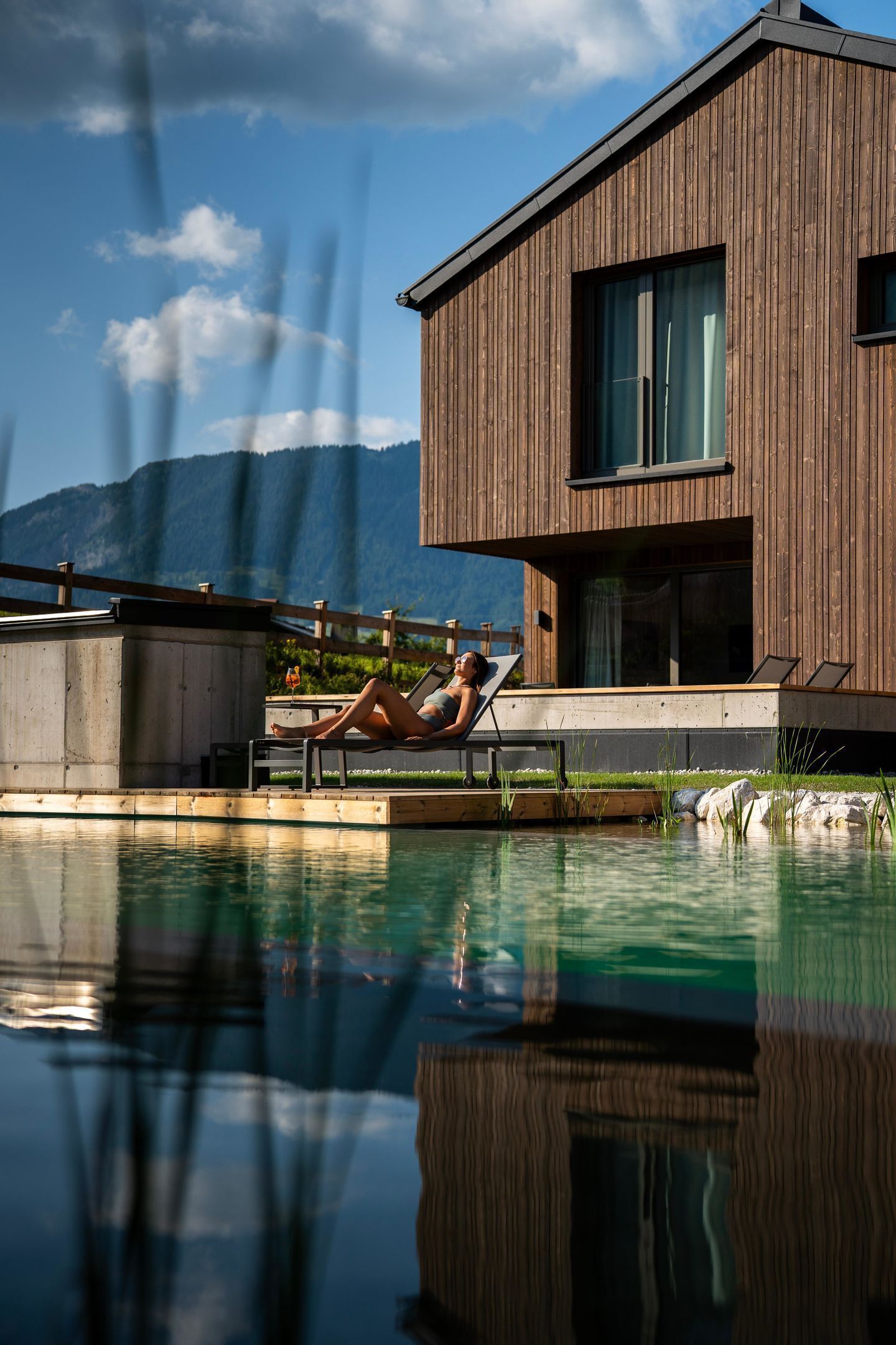 A modern house with a pool, a person relaxing on a deck, and mountains in the background.
