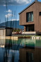 Una casa moderna con piscina, una persona relajándose en la terraza y montañas al fondo.