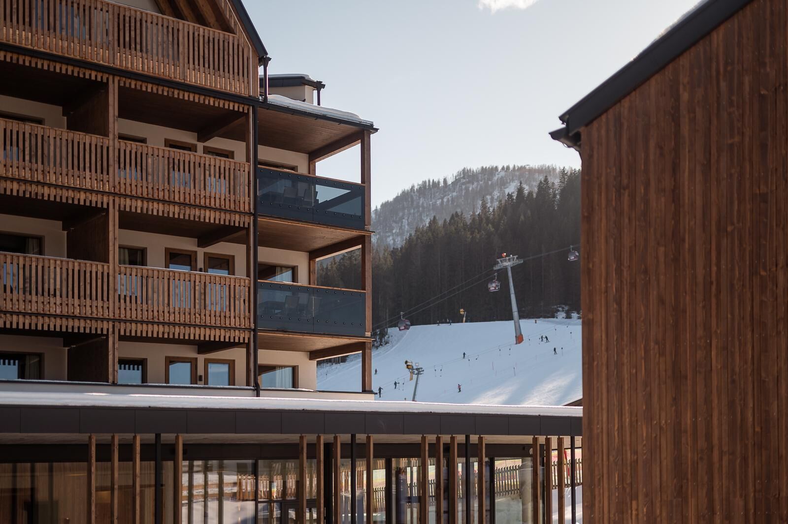 Un edificio de madera de varios pisos con balcones, una pista de esquí al fondo y un telesilla.