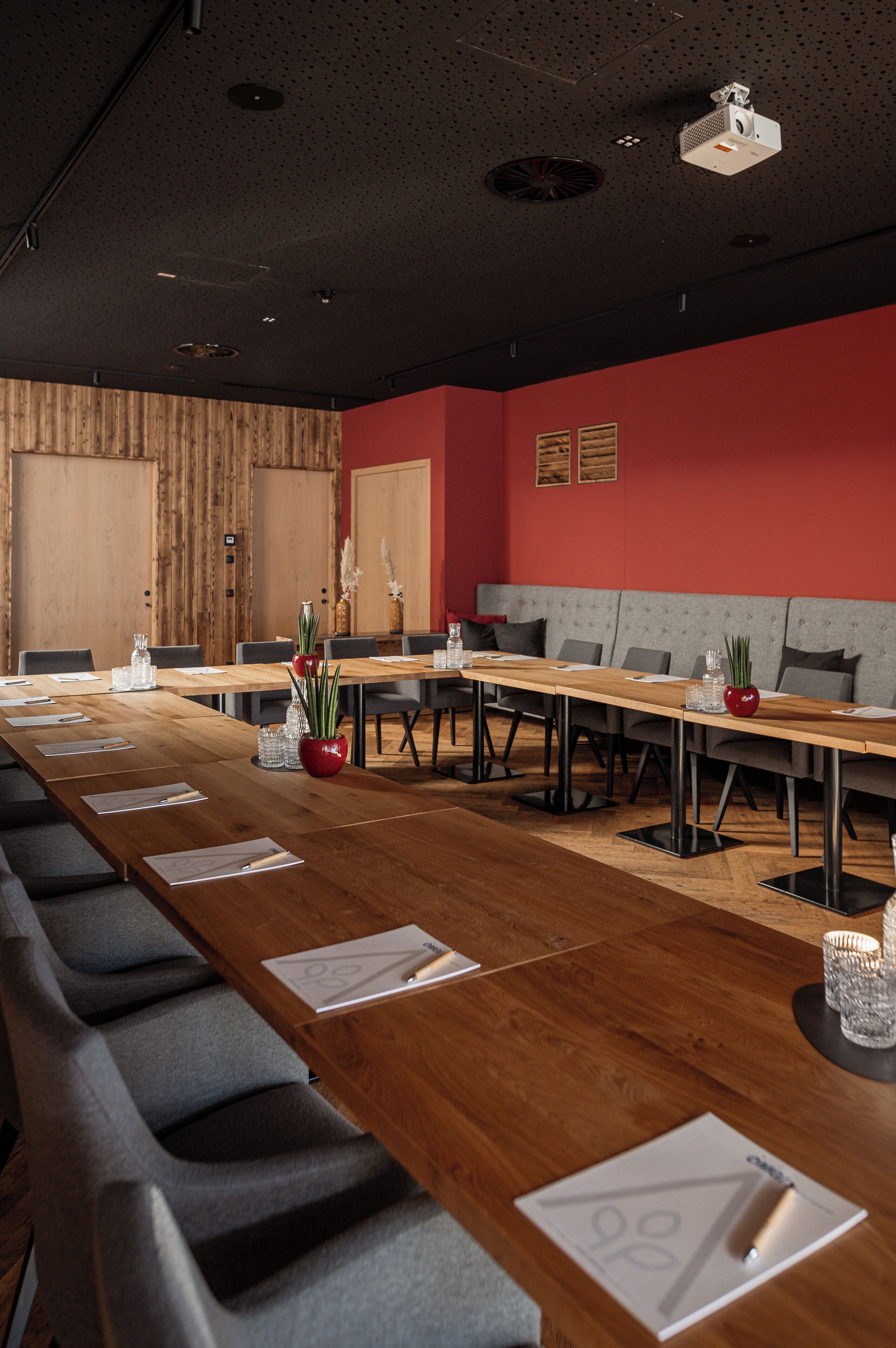 A conference room with a long wooden table, grey upholstered chairs, and a red accent wall.