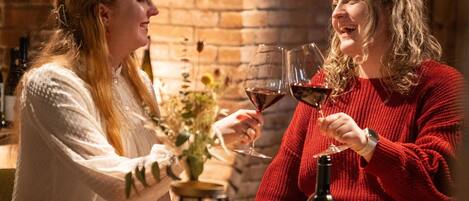 Two people toasting with wine glasses at a wooden table in a brick-walled bar area with shelves of wine bottles in the background.