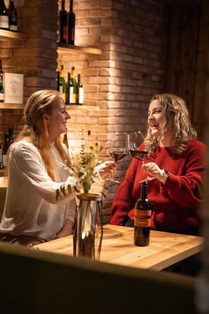 Dos personas brindando con copas de vino en una mesa de madera en un área de bar con paredes de ladrillo y estantes llenos de botellas de vino al fondo.